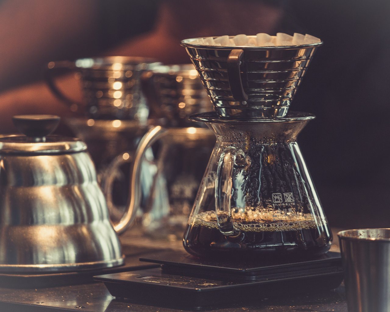 A steel pour-over dripper on a glass server on a scale, a gooseneck kettle beside it on a dark bar