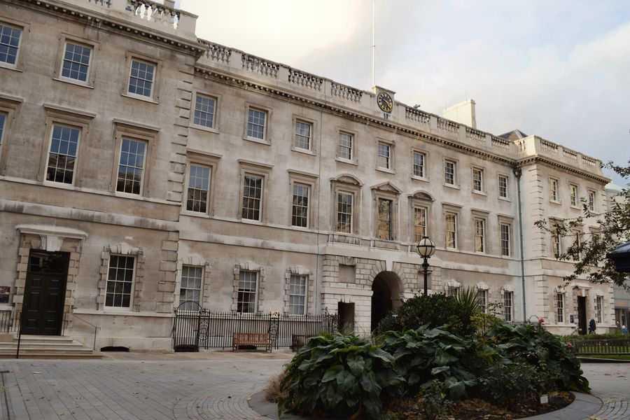 Pale stone hospital wing seen across a paved courtyard, with a large shrub bed in the foreground