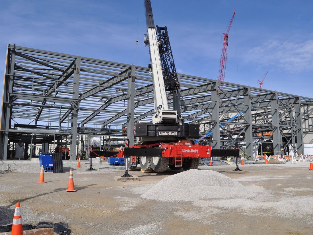 A long, low steel portal frame going up, with a red-and-white mobile crane and traffic cones on the slab in front