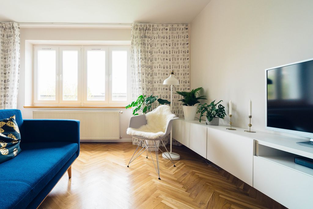 A bright living room: a blue sofa, white low cabinets, plants on the sill and a herringbone wooden floor