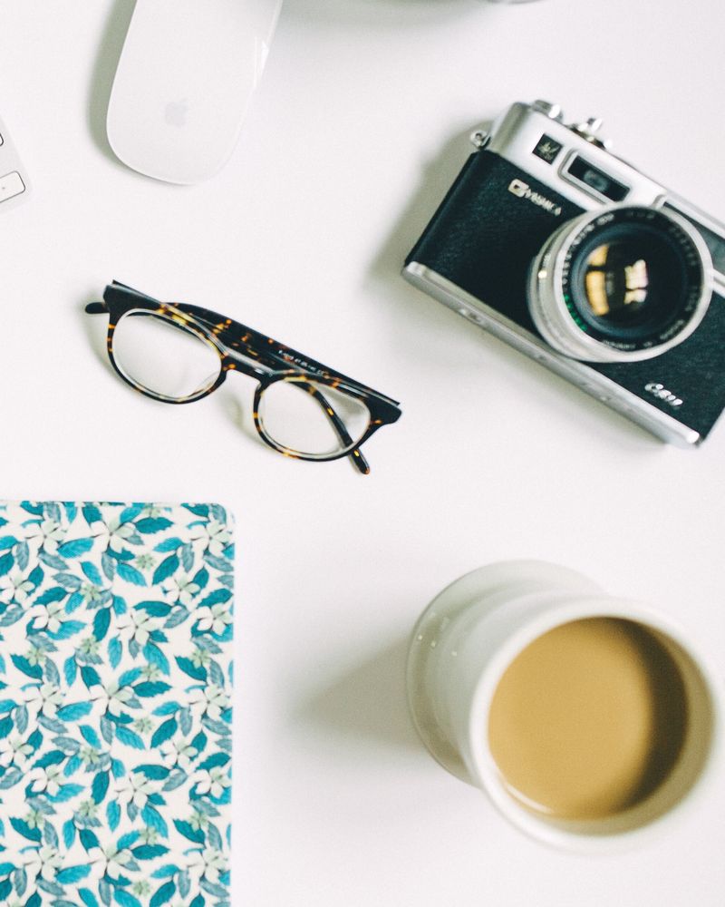 A desk from above with a camera, glasses, a mouse, a patterned notebook and a coffee