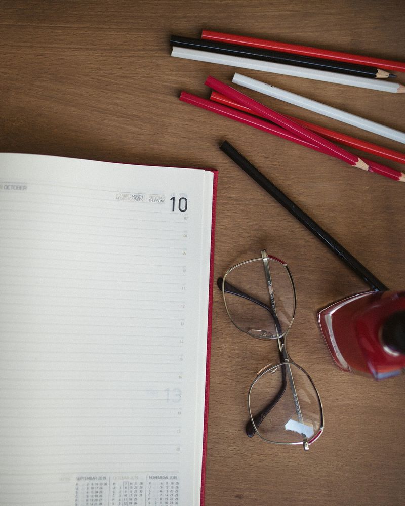 A diary open on a wooden desk beside red pencils and a pair of glasses
