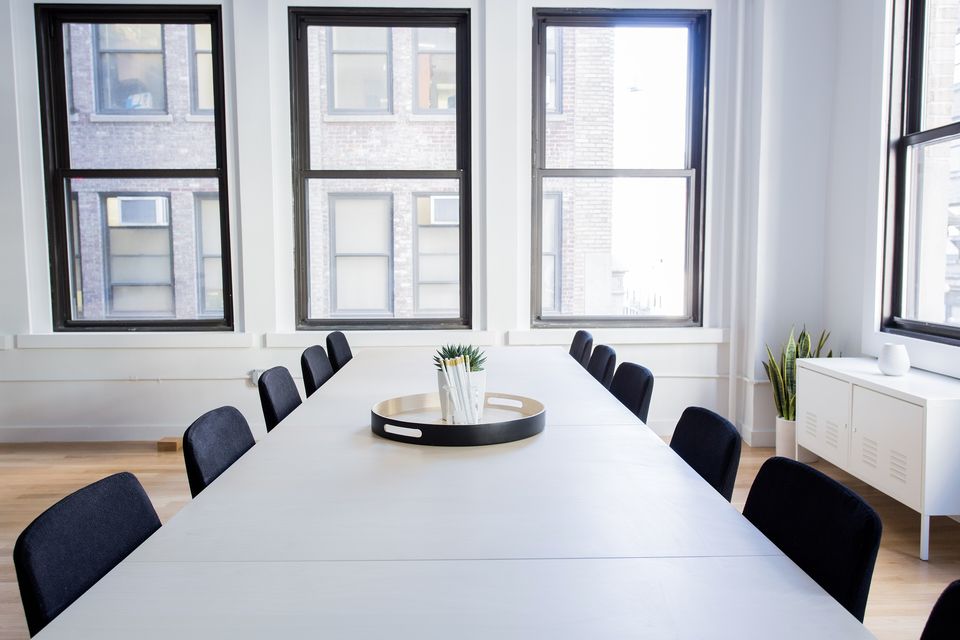 An empty meeting room with a long white table, black chairs and tall windows
