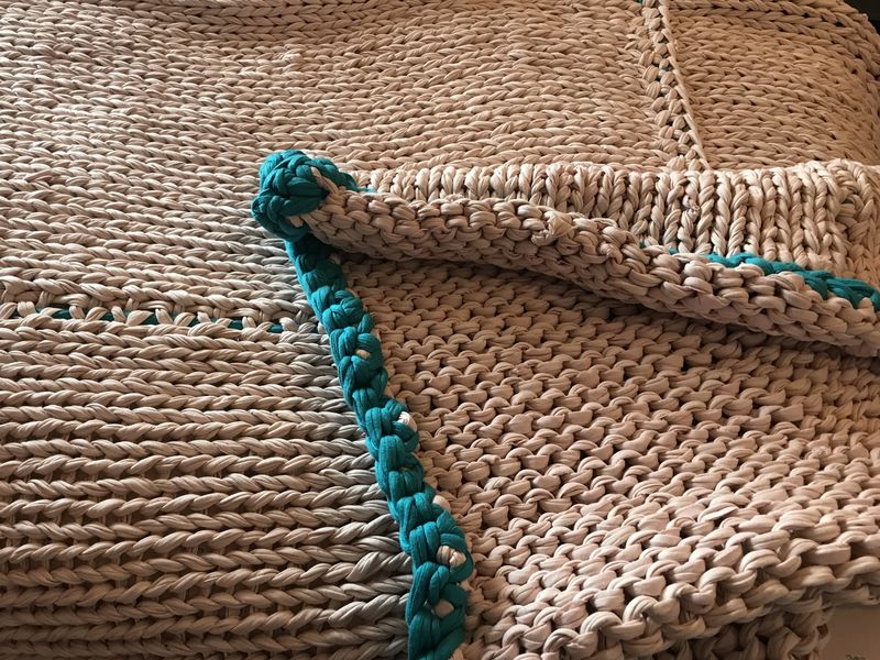 A thick knitted blanket in pale brown yarn with a teal edge, folded over