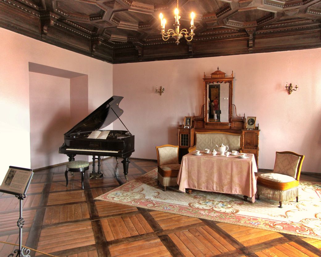 A salon with a grand piano, a carved wooden ceiling and a chandelier