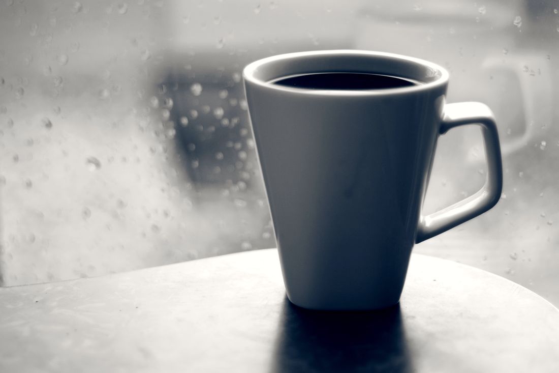 A grey mug of black coffee on a table by a rain-streaked window