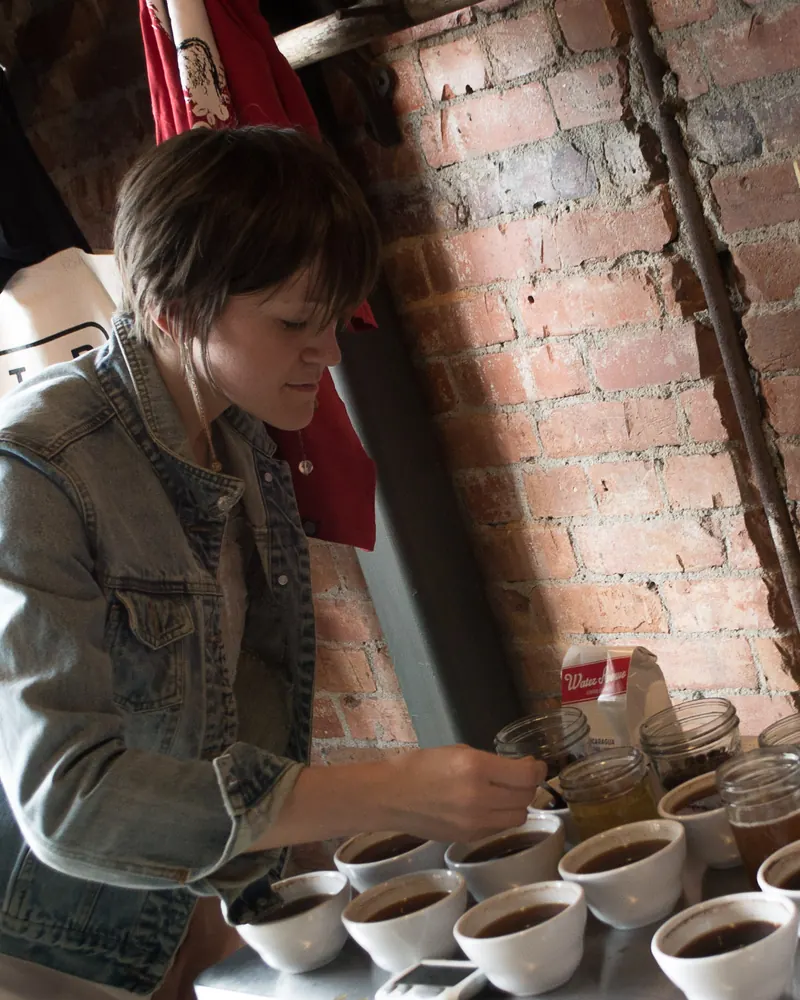 A taster at a cupping table, lifting a spoon of coffee beside rows of small white cups.
