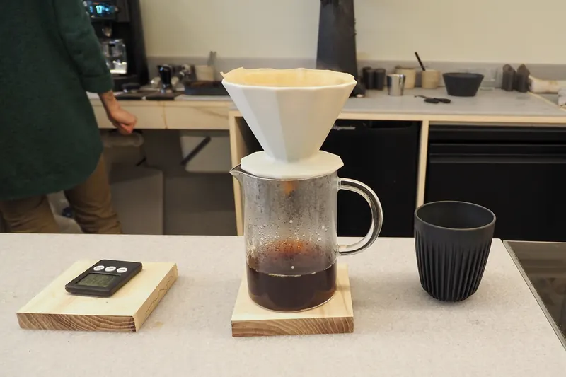A ceramic pour-over dripper on a glass jug of coffee, with a scale and a dark cup on a pale counter.