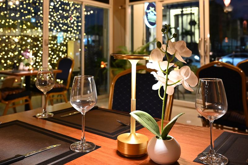 A restaurant table by the window at night, a small lamp and orchid on the table and fairy lights outside