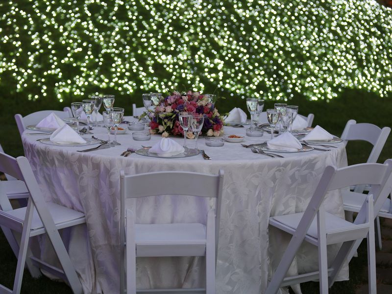 A round reception table laid with white linen and flowers, white folding chairs, a hedge of fairy lights behind