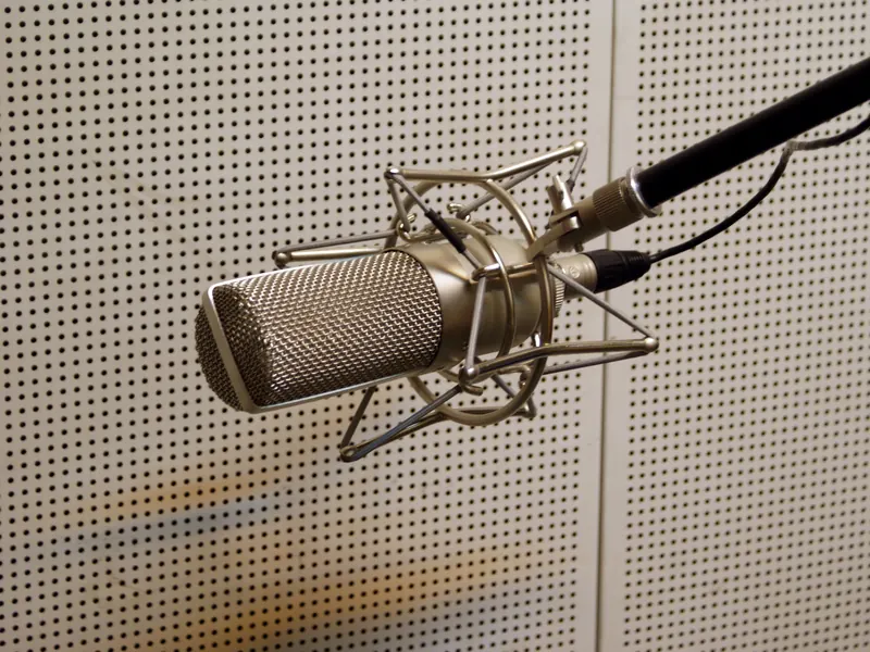 A ribbon microphone in a shock mount, hanging from a boom in front of a perforated studio wall