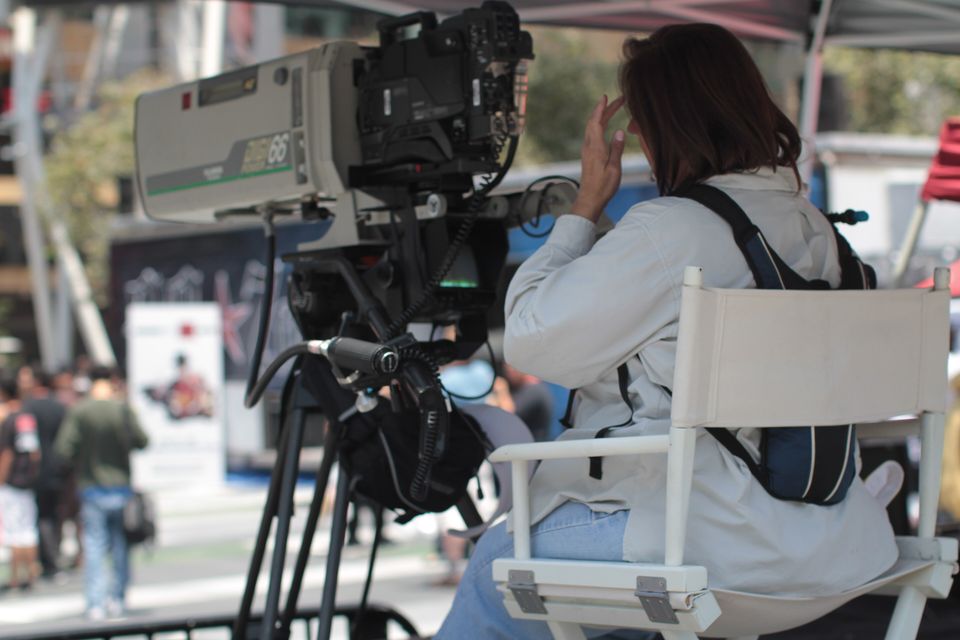 A television camera on a tripod at an outdoor event, with its operator seated beside it