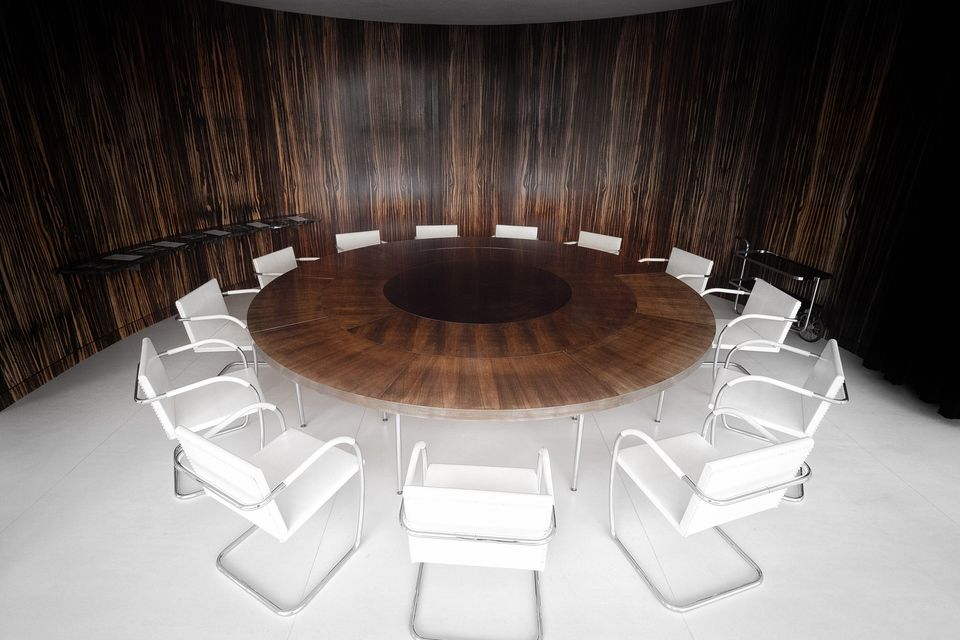 A round table and white chairs under a single lamp in a curved, wood-panelled room