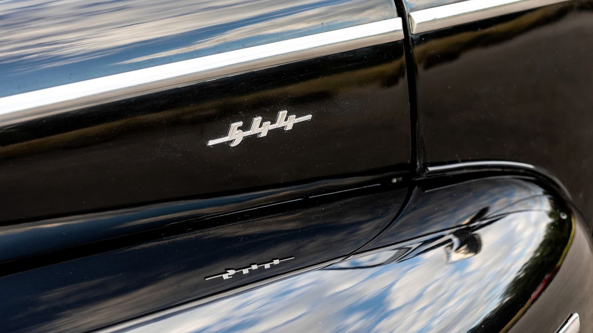 The flank of a gloss black classic car, with sky and trees reflected in the paint