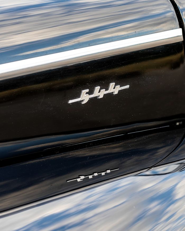 The flank of a gloss black classic car, with sky and trees reflected in the paint