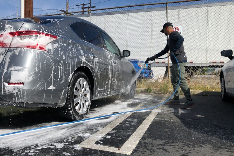A worker foaming a silver car with a hand sprayer in an open car park