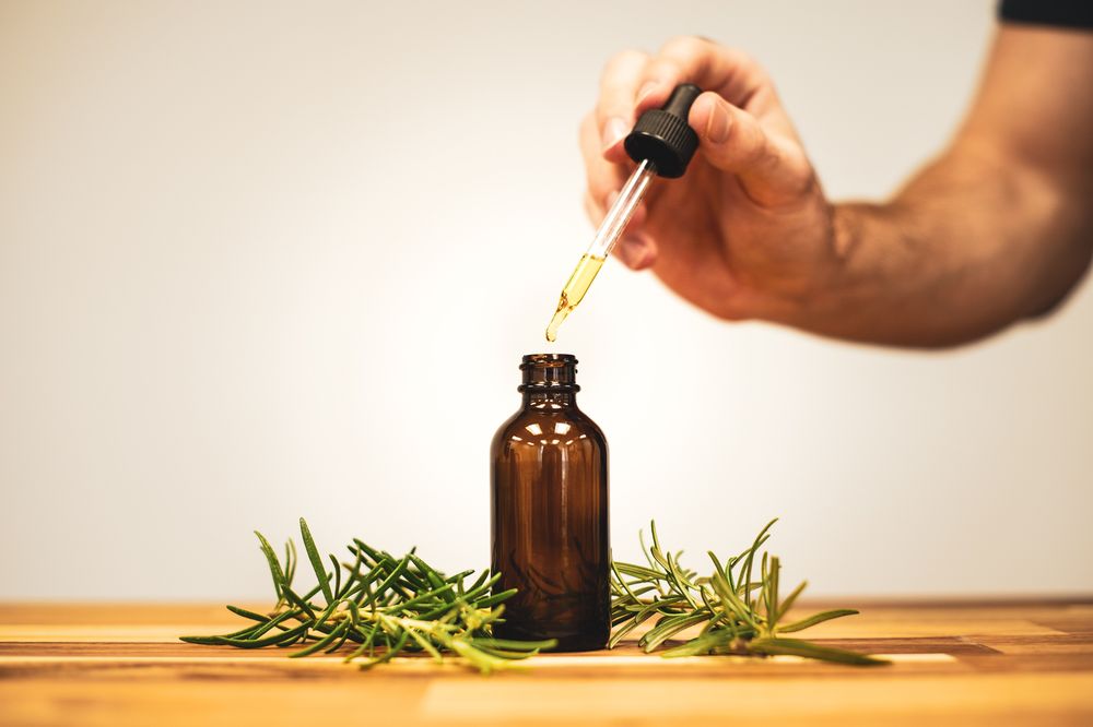 A hand holding a glass dropper above an amber bottle beside sprigs of rosemary