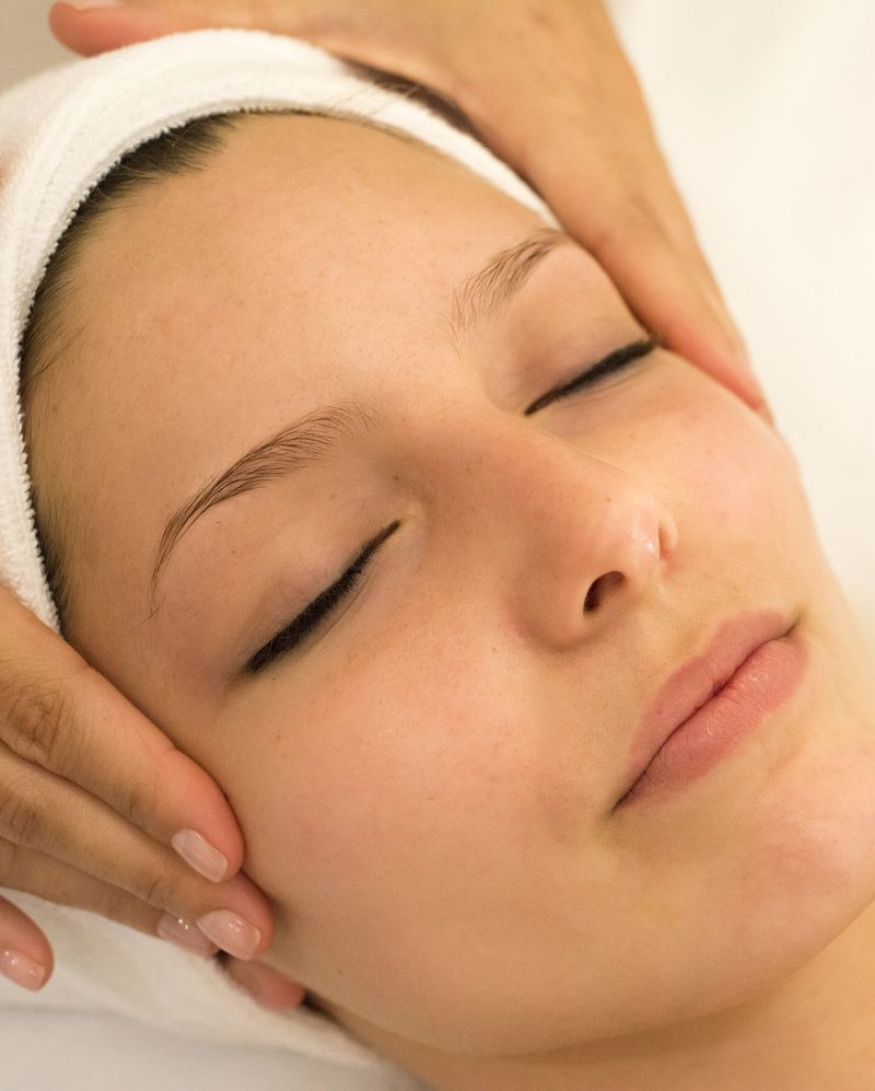 A facialist’s hands resting at a client’s temples as she lies back in a white headband, eyes closed