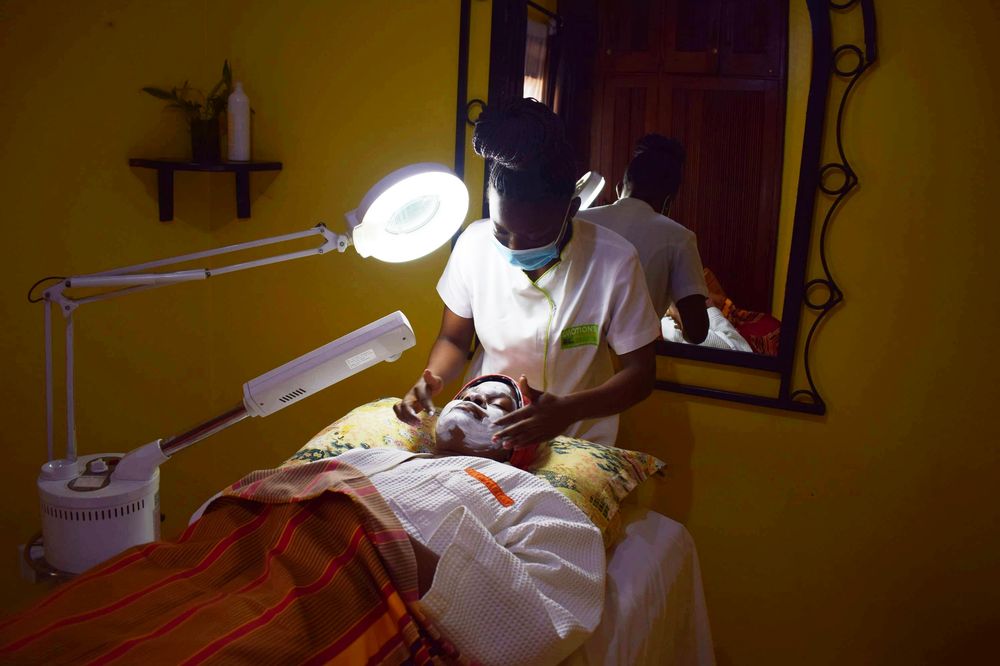 A facialist in a white tunic working on a client’s face under a magnifier lamp in a warm treatment room