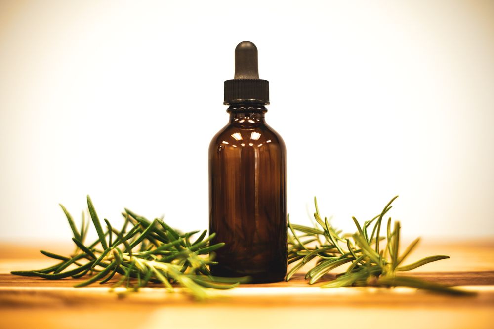 An amber dropper bottle between sprigs of rosemary on a pale wooden table