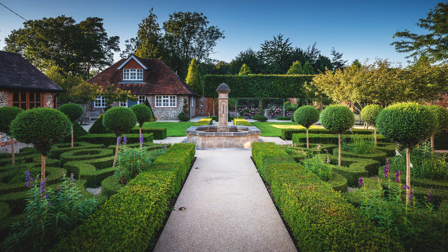A formal garden with clipped hedges, standard trees and a stone fountain in front of a tile-roofed house