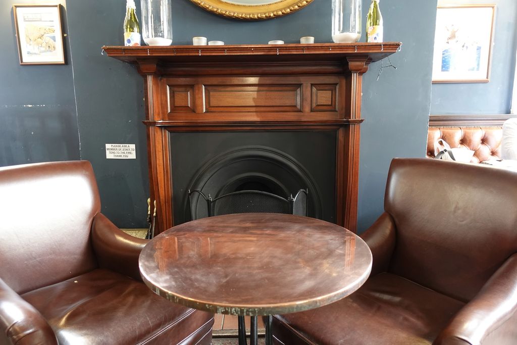 An open fireplace in a pub, with a round wooden table between two leather armchairs