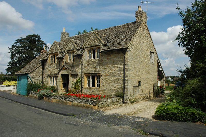 Honey-coloured Cotswold stone cottage with dormer windows beside a quiet lane