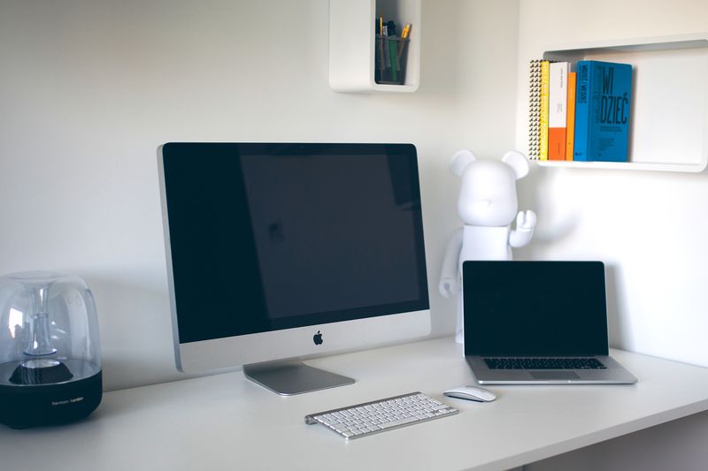 A tidy white desk with a desktop computer, a laptop and a small lamp