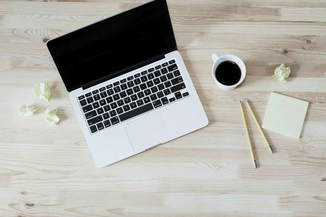 A laptop, a cup of black coffee, a sticky note and two pencils on a pale wooden desk, seen from above
