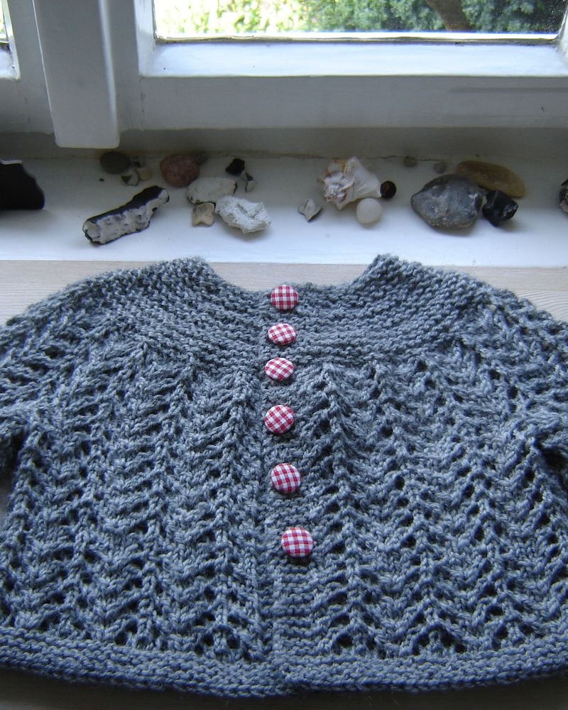 A grey hand-knitted baby cardigan with pink buttons, laid on a windowsill