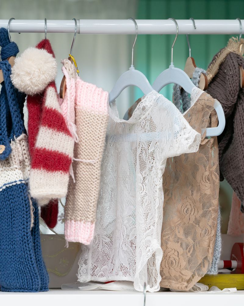 Small lace bodysuits and knitted baby clothes on hangers, with a teddy bear below
