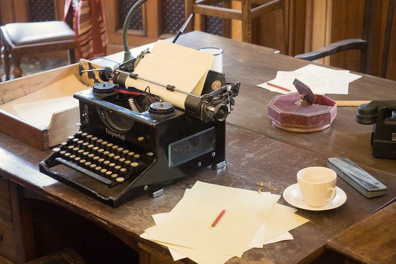A typewriter, loose paper and a cup on a worn wooden desk