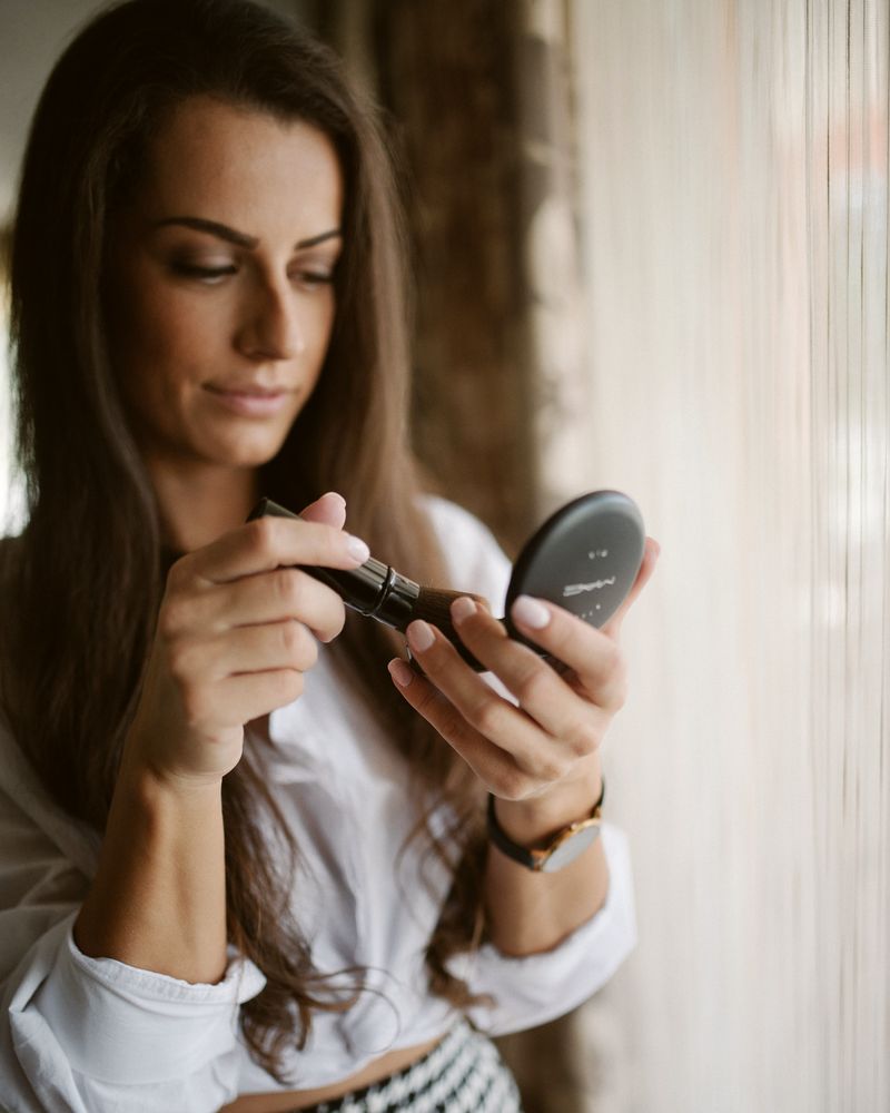 A woman by a tall window checking a small compact mirror, lit by the daylight