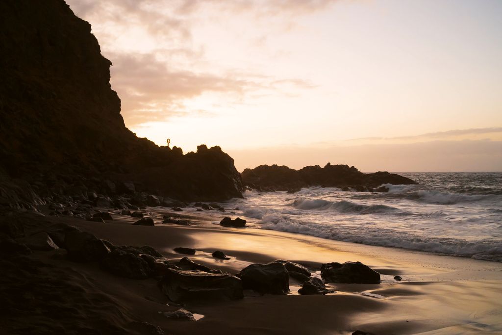 A dark rocky beach at dusk under a pink and orange sky, with sea stacks offshore