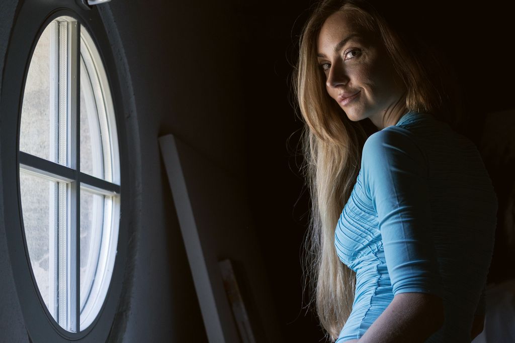 A woman in a blue top sits by a round porthole window in a dark room, lit only from the window