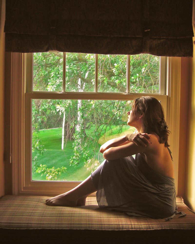 A woman sitting in a deep window seat, looking out at green trees, lit from the side