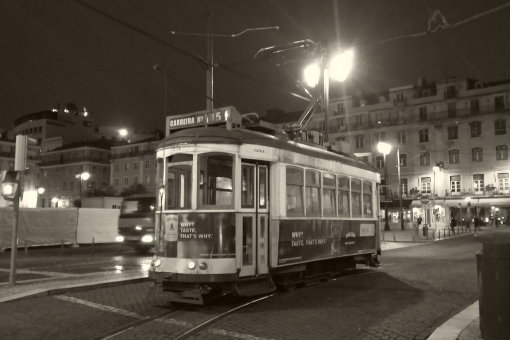 A tram on a dark Lisbon square at night under street lamps, in black and white