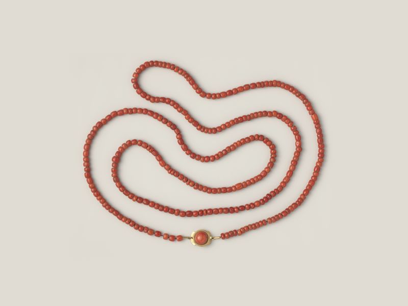 A necklace of strung red coral beads with a gold clasp, laid in loops, on pale grey, library photograph shown for style