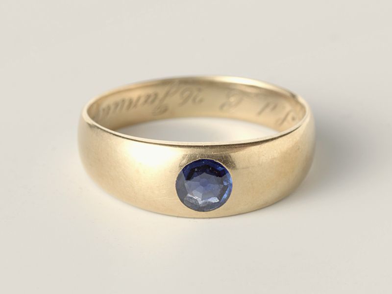 A smooth gold band set with a single small sapphire, on pale grey, library photograph shown for style
