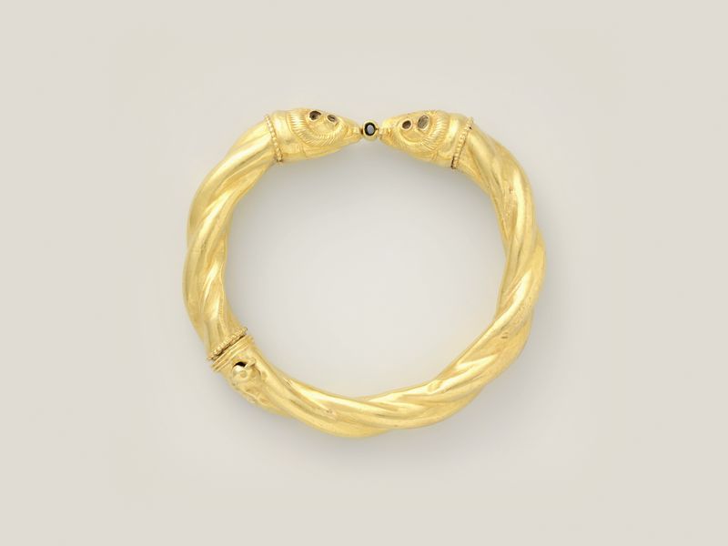A gold bangle of two twisted coils ending in ram heads, on pale grey, library photograph shown for style
