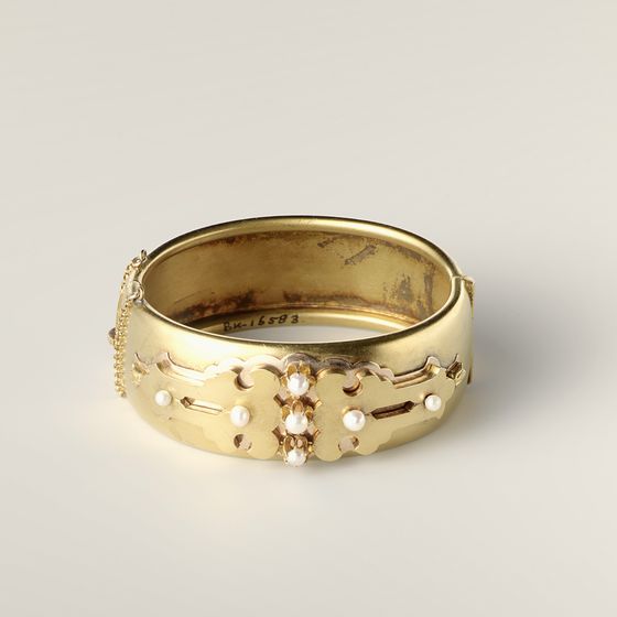A wide matt gold bangle with applied decoration and seven pearls, on pale grey, library photograph shown for style