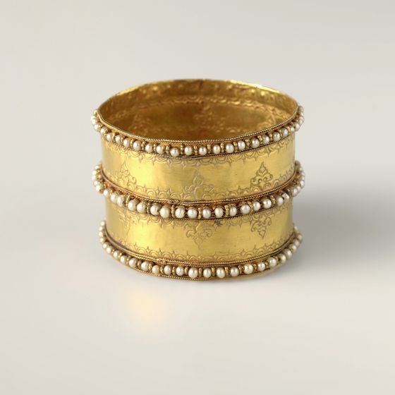 A wide gold cuff bracelet with three rows of seed pearls and engraving, on pale grey, library photograph shown for style