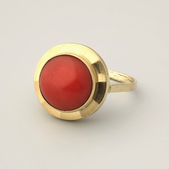 A gold ring with a single round red coral in a rub-over setting, on pale grey, library photograph shown for style