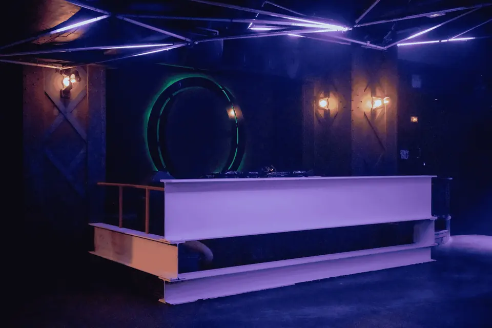 An empty white DJ booth in a dark club under blue light, with a lit ring on the wall behind