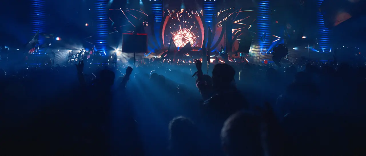 A crowd in silhouette facing a brightly lit stage through blue haze
