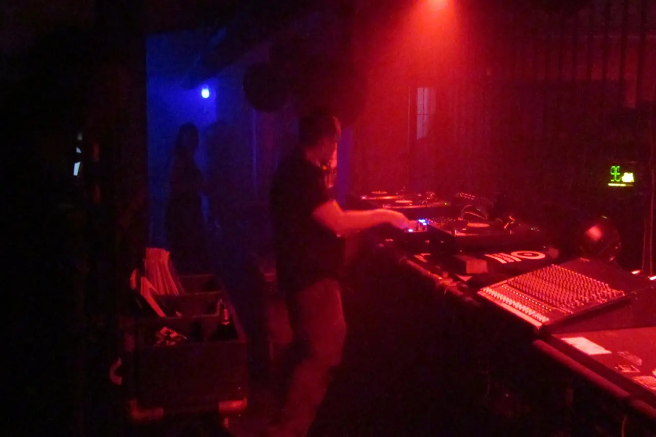 A DJ at the decks in a dark club booth lit red and blue, with a red spotlight overhead