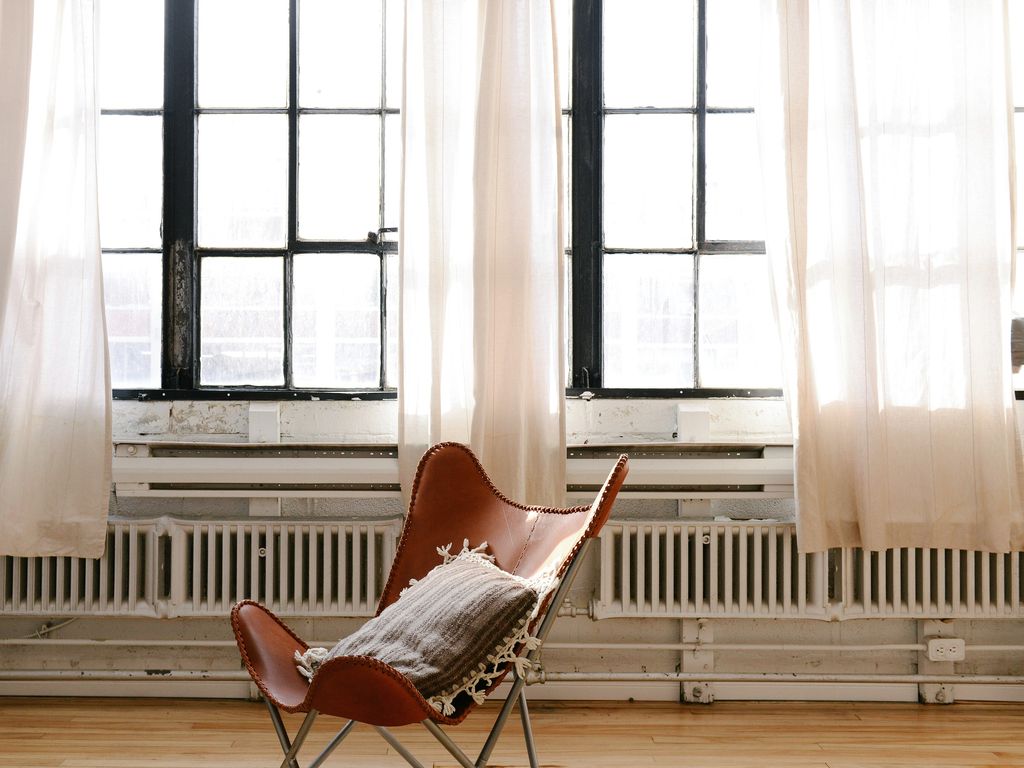 A leather sling chair in front of tall factory windows with sheer white curtains and a radiator
