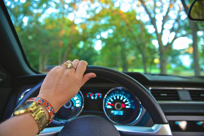 A driver’s hand resting on top of a steering wheel, with the dashboard dials below and trees through the windscreen.