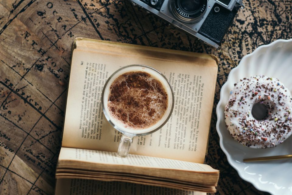 A cup of coffee and a doughnut beside an open book, seen from above