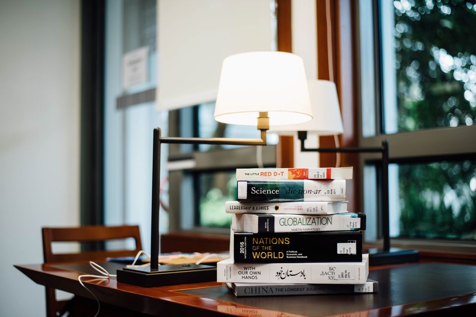 A stack of books under a desk lamp beside a window